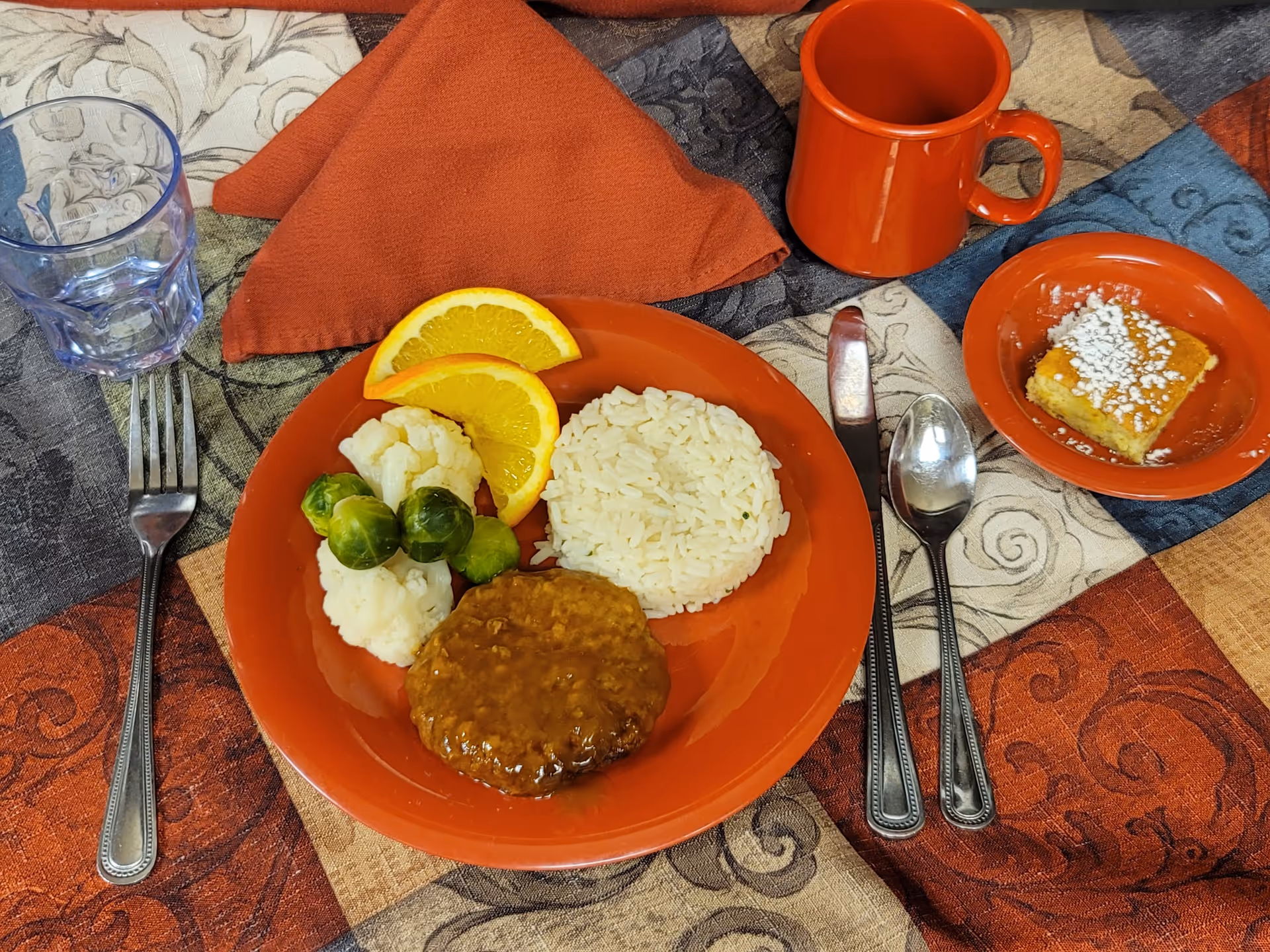 A meal served on an orange plate with white rice, a hamburger patty with gravy, cauliflower, Brussels sprouts, and two orange slices. Next to the plate is an orange mug, a small orange dish with a piece of cake topped with powdered sugar, a glass of water, and silverware on a multicolored patterned tablecloth with an orange napkin.