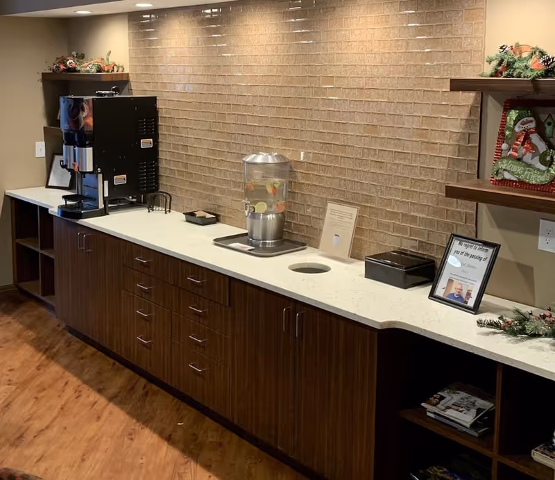 A countertop area with a beverage dispenser filled with fruit-infused water, a coffee machine, and a framed memorial photo with a tribute note. The countertop is white with dark wood cabinets below and a beige brick-patterned wall behind. There are shelves with holiday decorations and magazines on the right side.