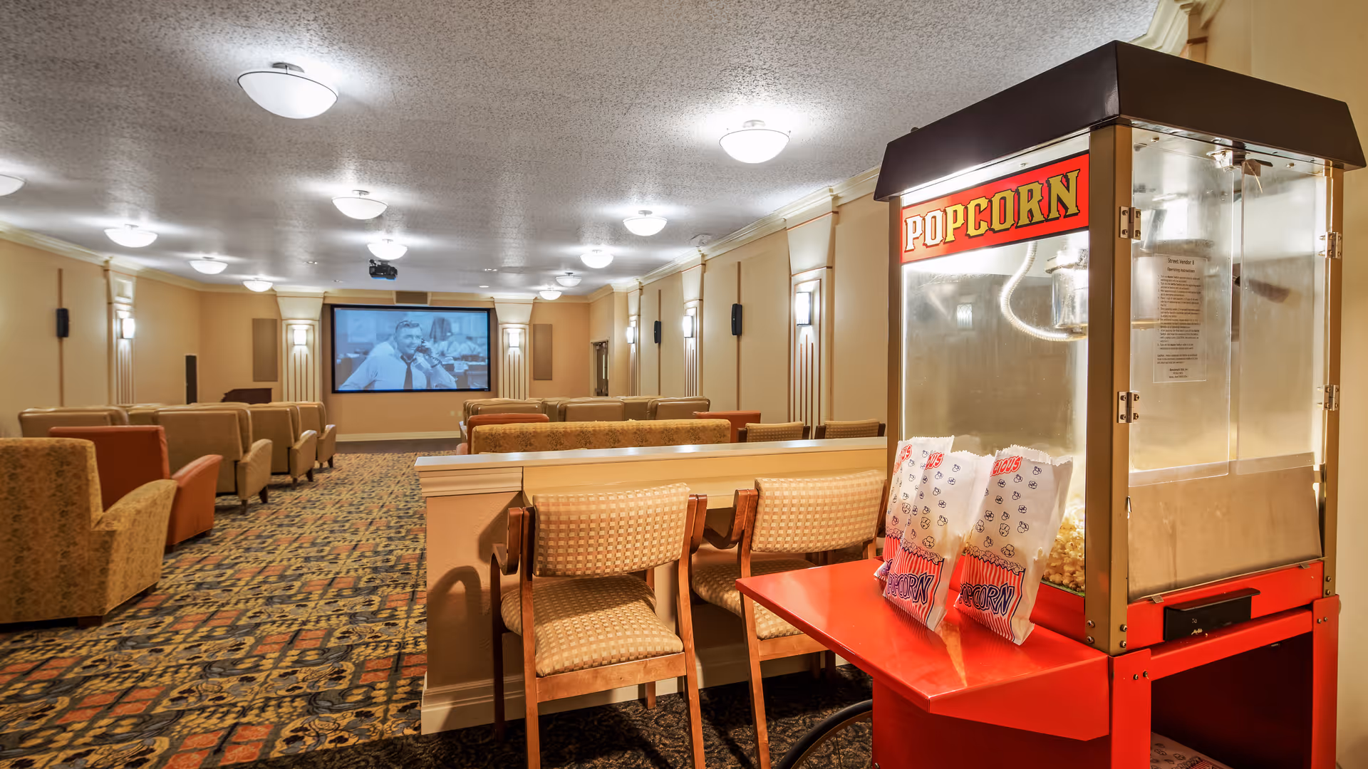 Interior view of a senior living facility's movie theater room with rows of cushioned chairs facing a large screen showing a black and white film. In the foreground, there is a popcorn machine with bags of popcorn on a red table attached to it. The room has patterned carpet and soft lighting from ceiling fixtures.