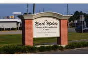 Outdoor view of a sign for North Mobile Nursing & Rehabilitation Center, located on a grassy area with a building and parked cars in the background under a clear sky.
