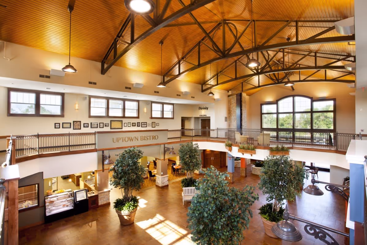 Spacious and well-lit interior of a senior living facility featuring a high wooden ceiling with exposed beams, large windows allowing natural light, potted plants, and a mezzanine level with railings. Below, there is a dining area labeled 'Uptown Bistro' with tables and chairs, and a sign indicating a chapel nearby.