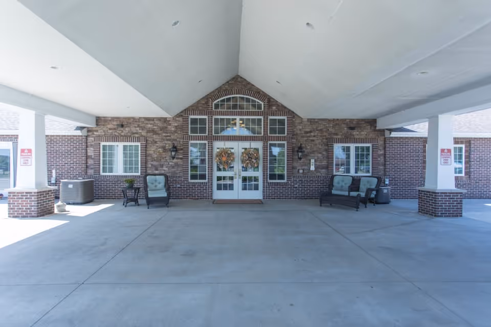 Covered driveway and entrance of a brick senior living building with double glass doors adorned with wreaths and seating on either side.