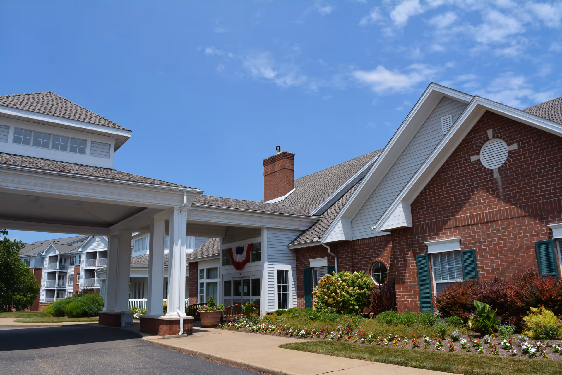 Exterior view of The Glenwood Community building with a covered entrance, brick and white siding walls, landscaped flower beds, and a clear blue sky.