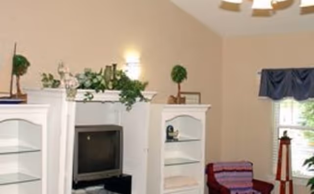 A cozy living room area with a white entertainment center holding a television and decorative plants on top. There are two white shelving units on either side of the TV. A window with a blue valance lets in natural light, and a comfortable chair with a colorful blanket is positioned near the window. The walls are painted beige, and there is a ceiling light fixture visible.