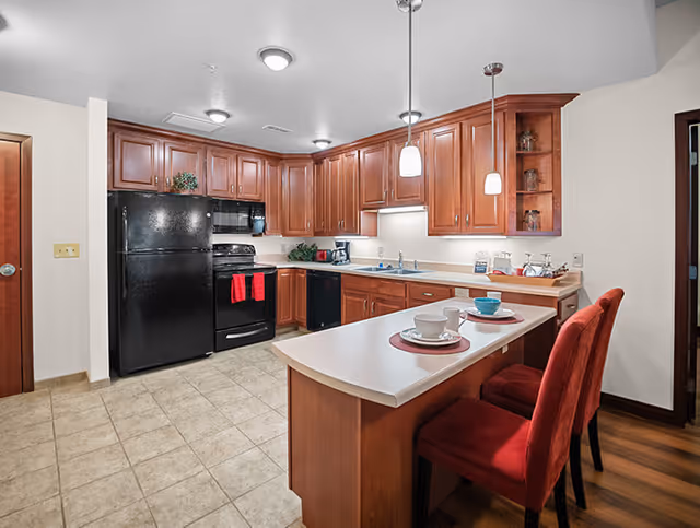 A modern kitchen with wooden cabinets, black refrigerator and stove, a white countertop island with two red cushioned chairs, and pendant lights hanging above the island. The kitchen has tiled flooring and is well-lit with ceiling lights.