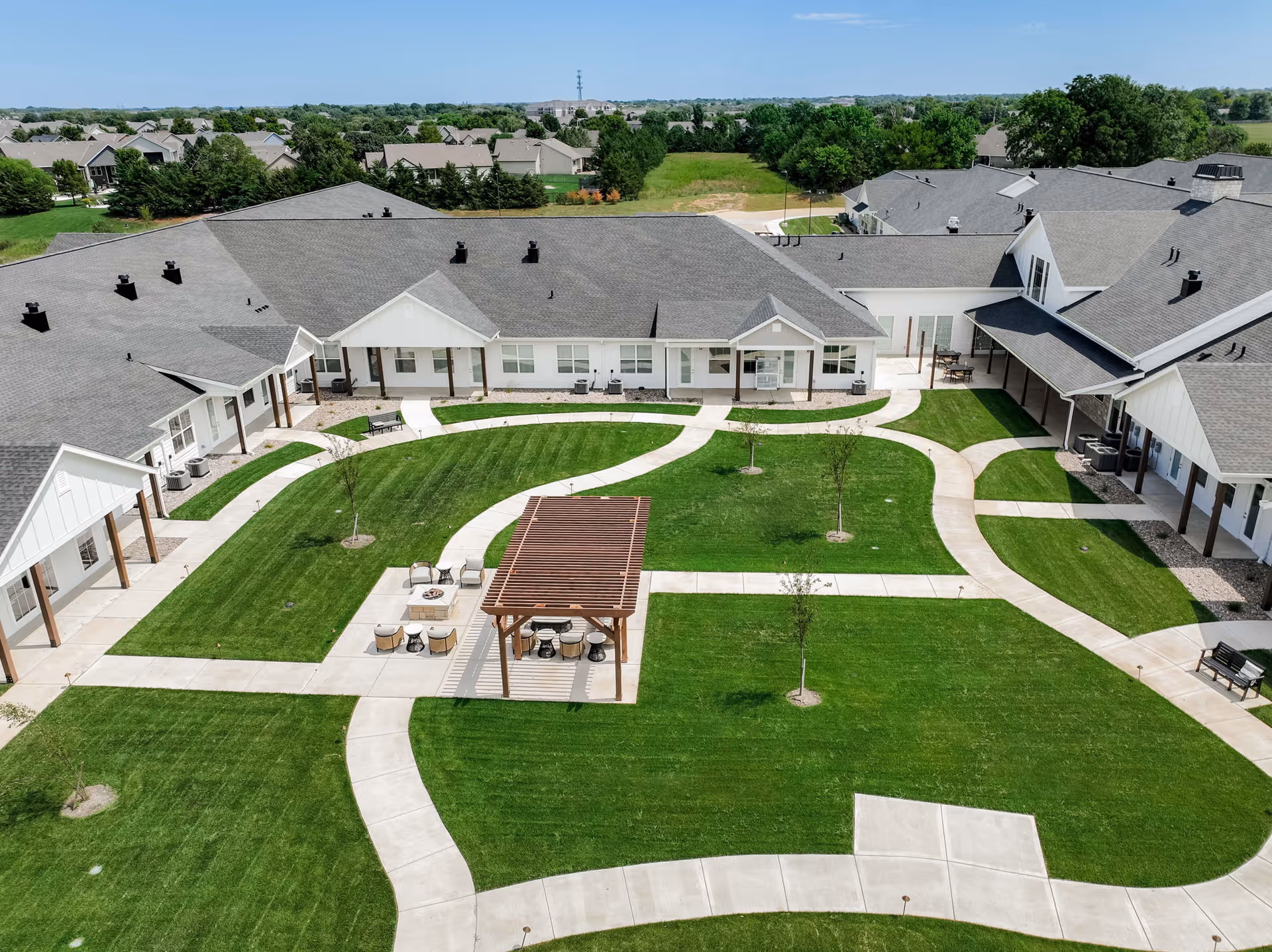 Aerial view of a senior living facility courtyard with well-maintained green lawns, paved walkways, small trees, and a wooden pergola with seating underneath. The surrounding buildings have gray roofs and white walls with covered walkways.