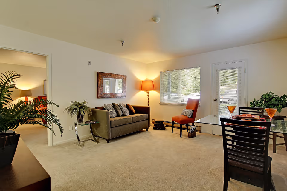 A cozy living room with a gray sofa adorned with multiple cushions, a tall floor lamp in the corner, a red chair with a small pillow, a glass dining table set with plates and glasses, and a window and door letting in natural light. There is a potted plant on a side table next to the sofa and another plant on a cabinet. An open doorway leads to a bedroom with a bed and lamp visible.
