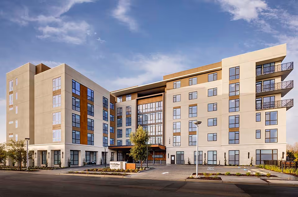 Exterior view of a modern multi-story senior living facility building with large windows, balconies, and a driveway entrance under a partly cloudy sky.