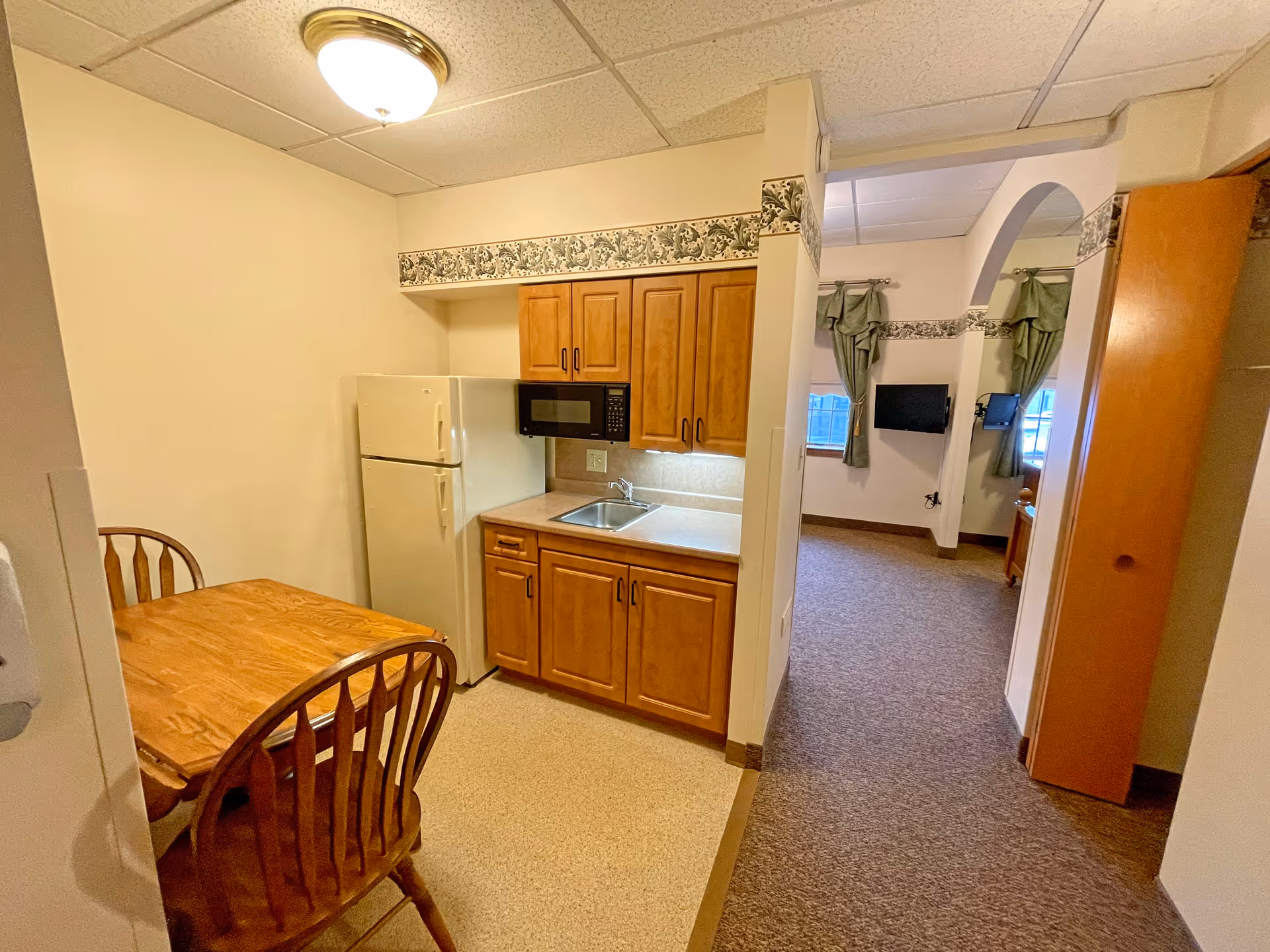 Small kitchenette area with wooden cabinets, a sink, a microwave, and a refrigerator. Next to the kitchenette is a wooden dining table with two chairs. The room has beige walls with a decorative floral border near the ceiling. In the background, there is an open doorway leading to a carpeted living area with windows covered by green curtains and a wall-mounted TV.