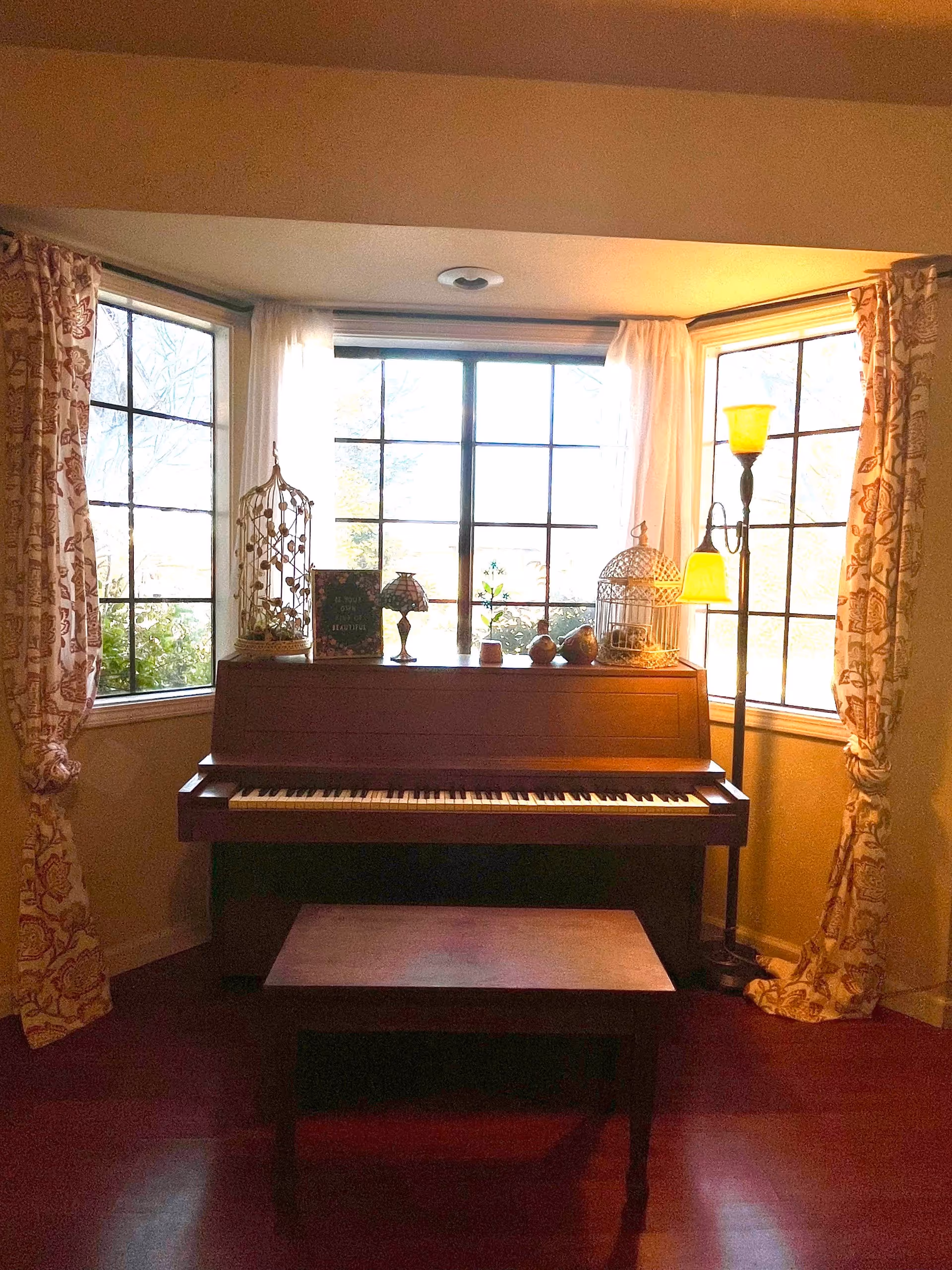 A wooden upright piano with a matching bench in front of a large bay window with three sections. The window is framed by patterned curtains and sheer white drapes. On top of the piano are decorative items including two birdcages, a small lamp, a book, and some small ornaments. To the right of the piano is a floor lamp with two yellow lampshades. The room has warm lighting and a cozy atmosphere.