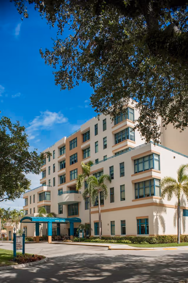 Exterior view of Hazel Cypen Tower, a multi-story building with beige and white walls and teal window frames. The entrance has a teal canopy with the building name displayed. Palm trees and other greenery surround the building under a clear blue sky.