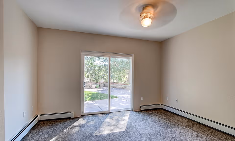 Empty room with beige walls and carpeted floor, featuring a ceiling light fixture and a large sliding glass door that opens to an outdoor patio area with greenery.