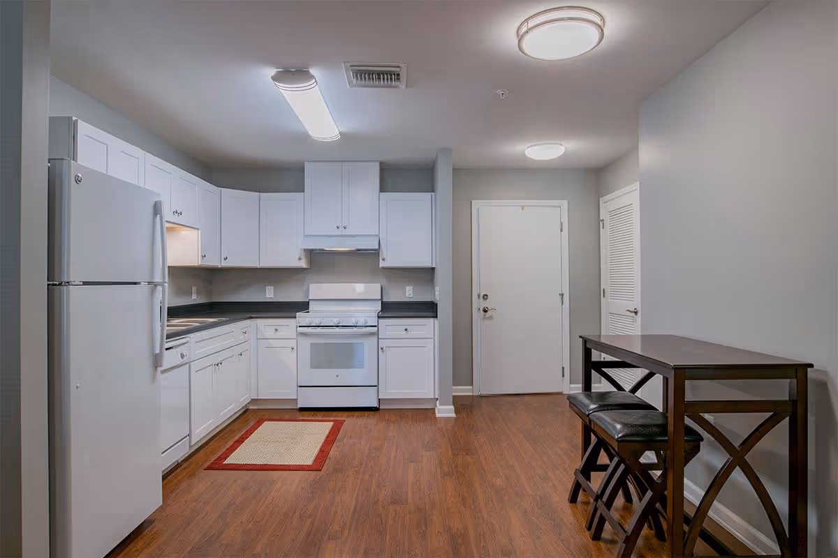 A modern kitchen with white cabinets, a white refrigerator, a white stove with an overhead vent, and a dishwasher. The kitchen has dark countertops and wood flooring. To the right, there is a small dining area with a dark wooden table and two matching stools. The walls are painted light gray, and there are ceiling lights providing illumination.
