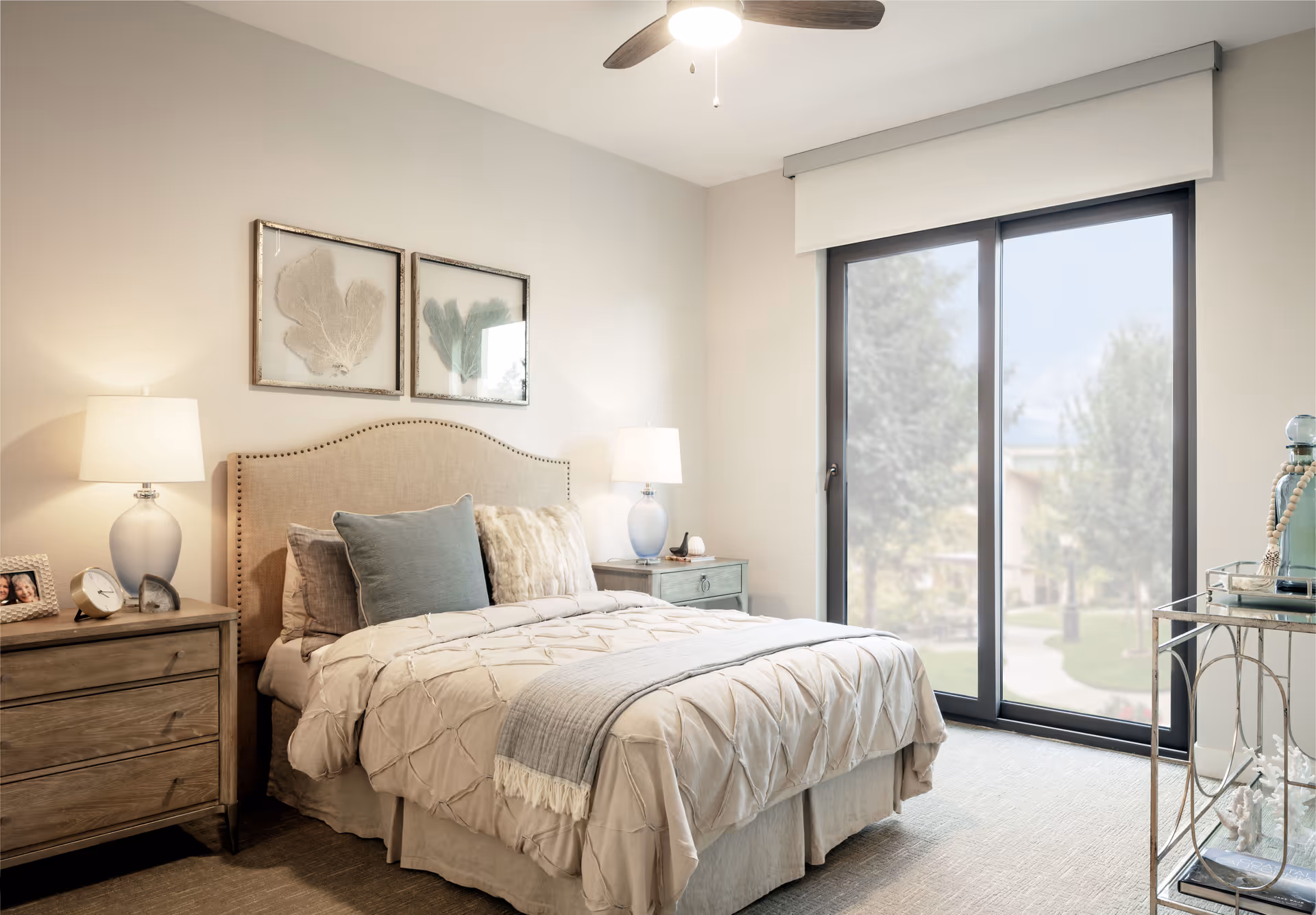 A cozy bedroom with a beige upholstered bed adorned with multiple pillows and a textured comforter. Two wooden nightstands with lamps flank the bed. Above the bed are two framed botanical prints. A large sliding glass door lets in natural light and offers a view of trees and a pathway outside. A ceiling fan with a light is mounted on the ceiling.