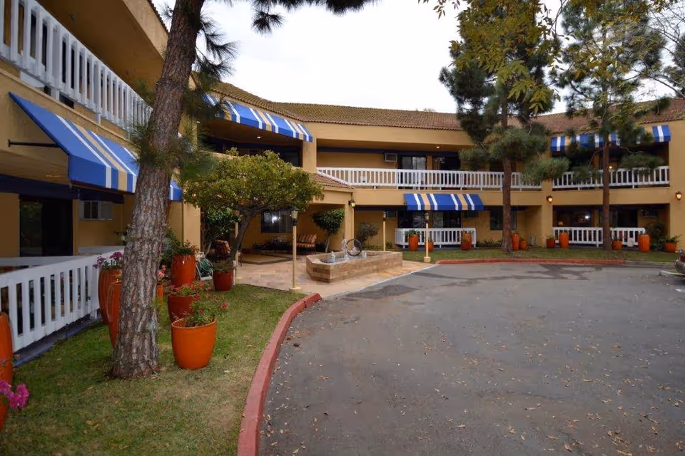 Two-story yellow U-shaped building with blue-and-white striped awnings, potted plants, trees and a circular driveway opening onto a central courtyard.