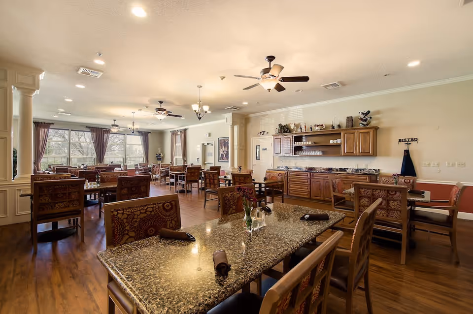 A spacious senior living dining room with multiple wooden tables and chairs, granite tabletops, ceiling fans, and large windows with curtains allowing natural light. The room features a wooden cabinet and countertop area with shelves holding glasses and decorative items.