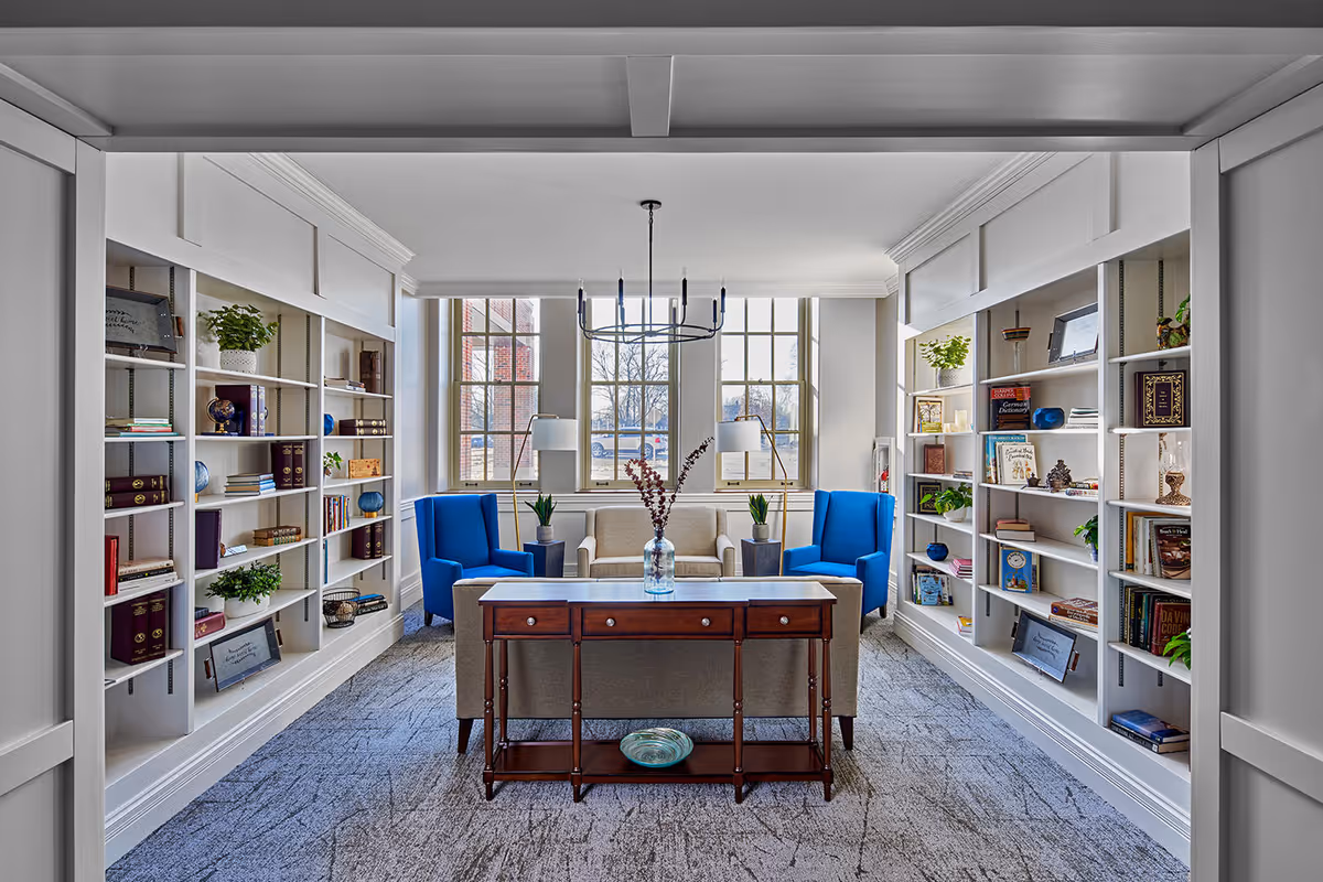 Bright communal library-style living room with built-in bookshelves, two blue armchairs, a sofa, and a central wooden console table in front of tall windows.