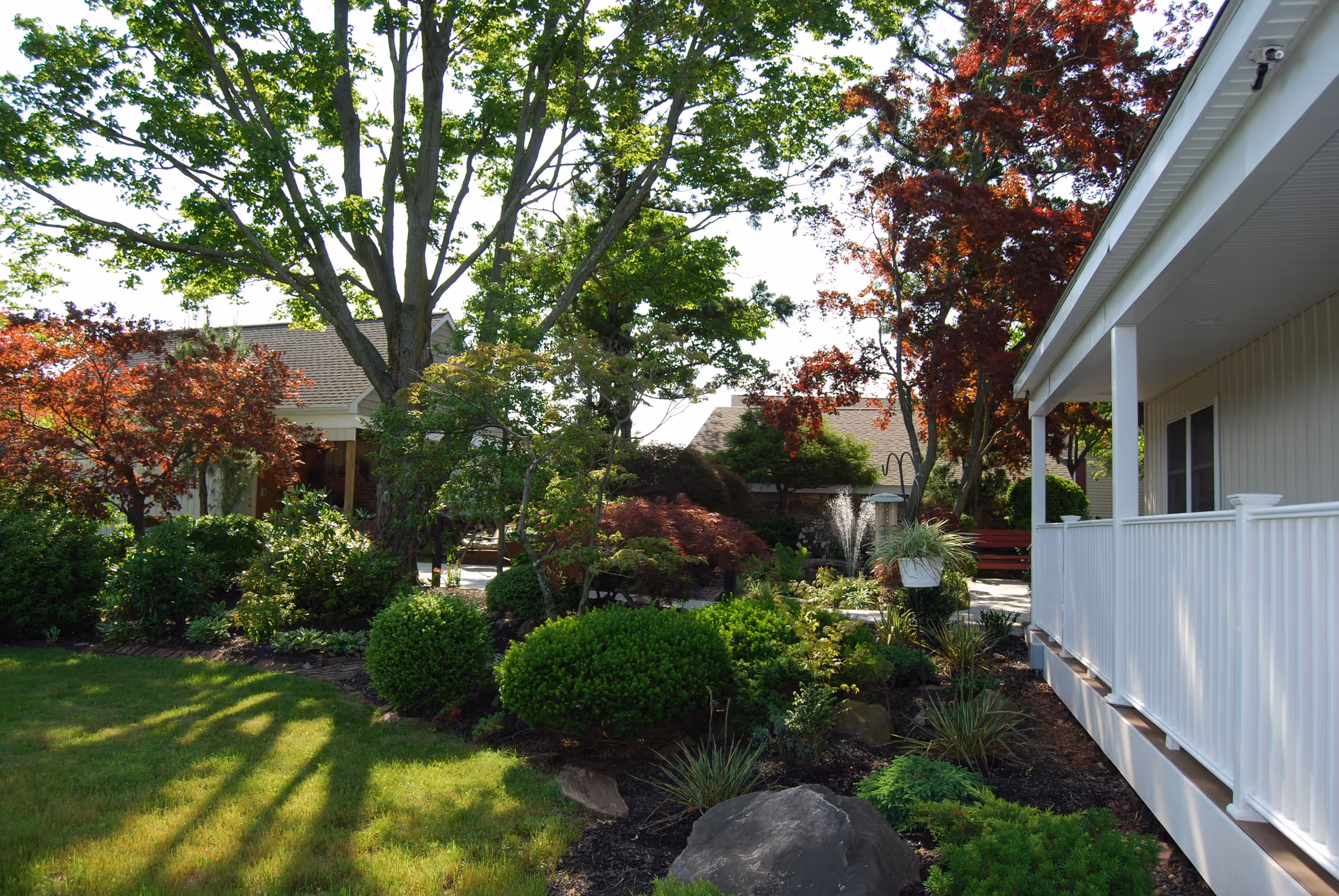 A landscaped garden area with various green shrubs, trees with green and red leaves, and a small water fountain. To the right, there is a white building with a porch and railing. The scene is bright and sunny with shadows cast on the grass.
