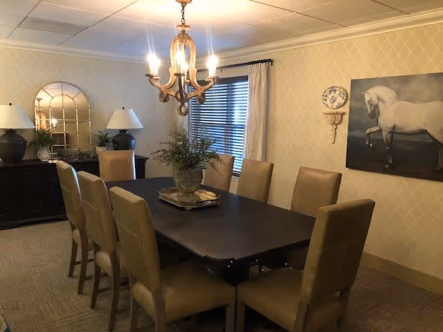 A formal dining room with a long dark wooden table surrounded by eight beige upholstered chairs. A chandelier with lit bulbs hangs above the table. The walls are decorated with patterned wallpaper, a large painting of a white horse, a decorative plate, and a mirror above a black sideboard with two lamps and a plant.