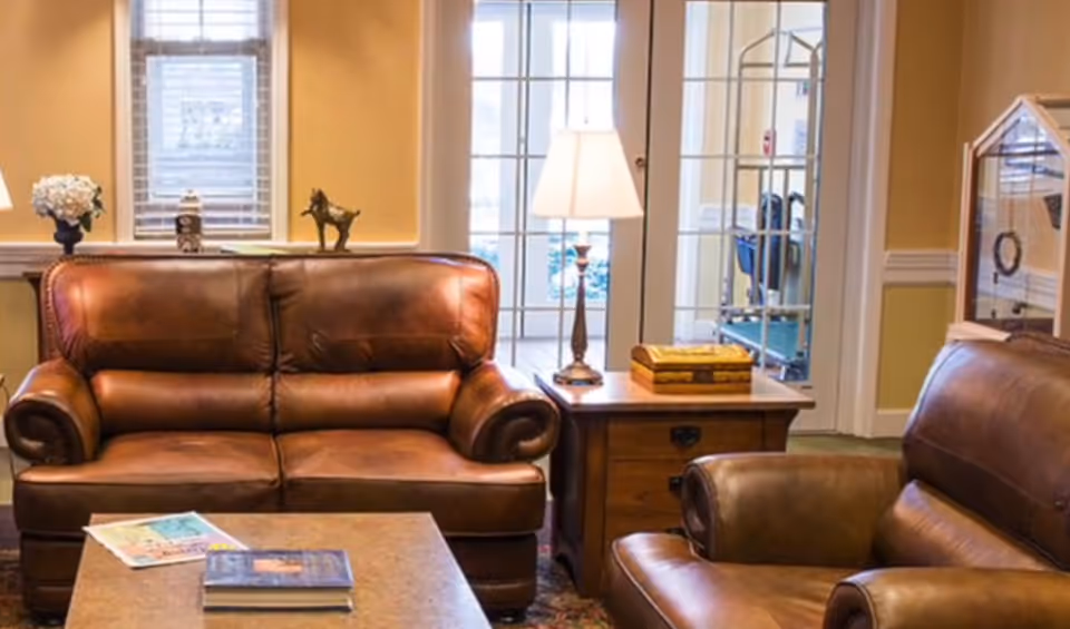 A cozy living room area with two brown leather sofas facing a coffee table. Behind one sofa is a window with white blinds and decorative items on the windowsill. A wooden side table with a lamp and a decorative box is placed between the sofas. In the background, there are glass doors leading to another room with exercise equipment visible.
