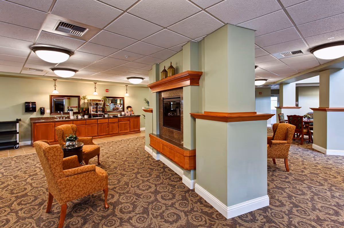 Interior view of a senior living community common area featuring a double-sided fireplace with wooden trim, two upholstered armchairs with a small round table between them, a carpeted floor with a patterned design, and a counter area with coffee and snack stations along the back wall.