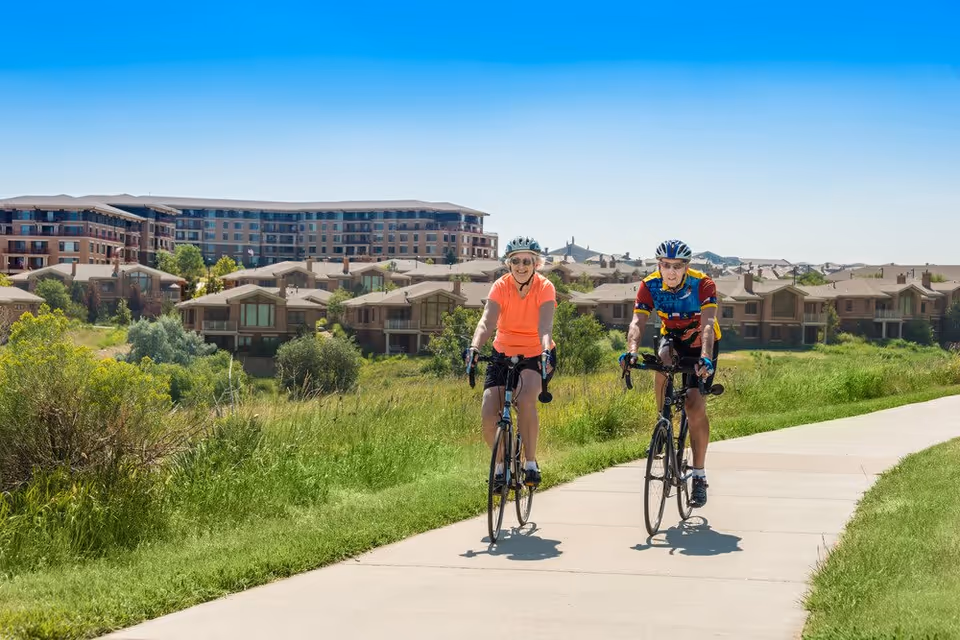 Two elderly people wearing helmets and cycling on a paved path surrounded by green grass and bushes, with residential buildings and a large apartment complex in the background under a clear blue sky.