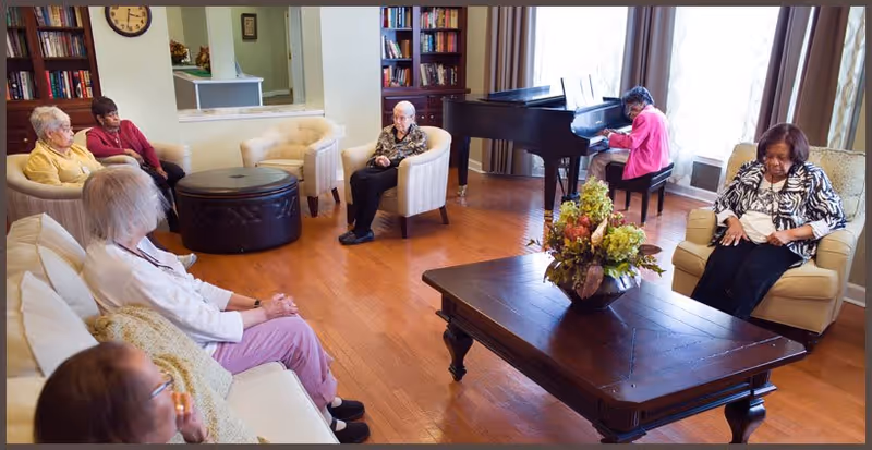 A group of elderly women seated in a cozy living room with wooden floors, cream-colored armchairs, a large wooden coffee table with a floral centerpiece, and a woman playing a grand piano near large windows with curtains.