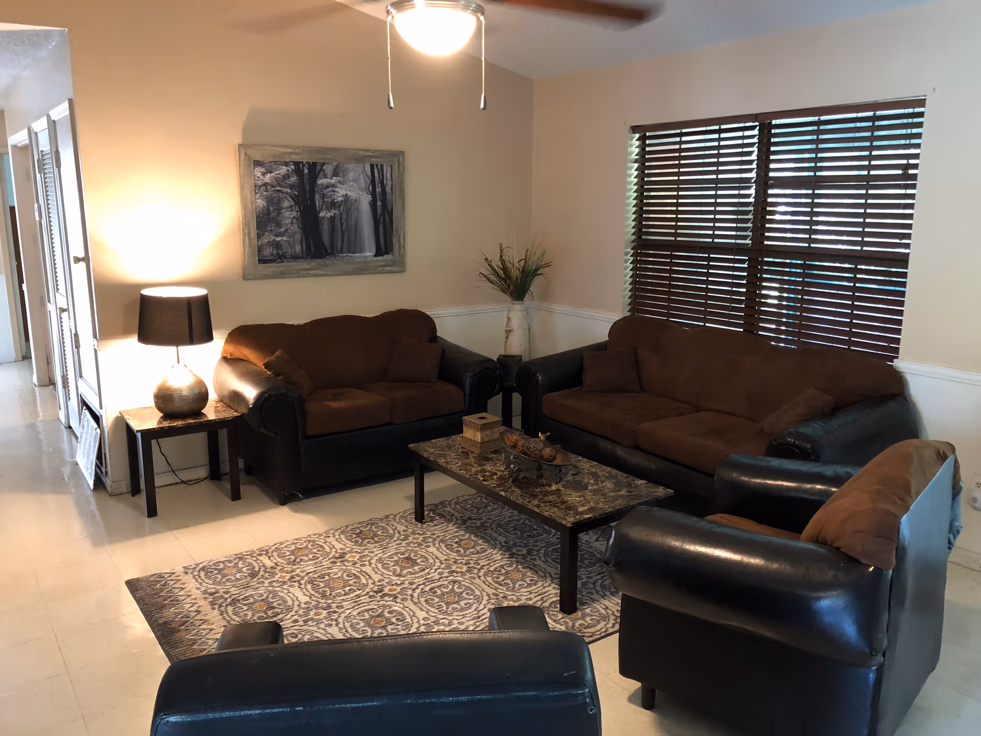 Cozy living room with brown sofas and armchairs arranged around a coffee table on a patterned rug, with a lamp, wall art, and window blinds.