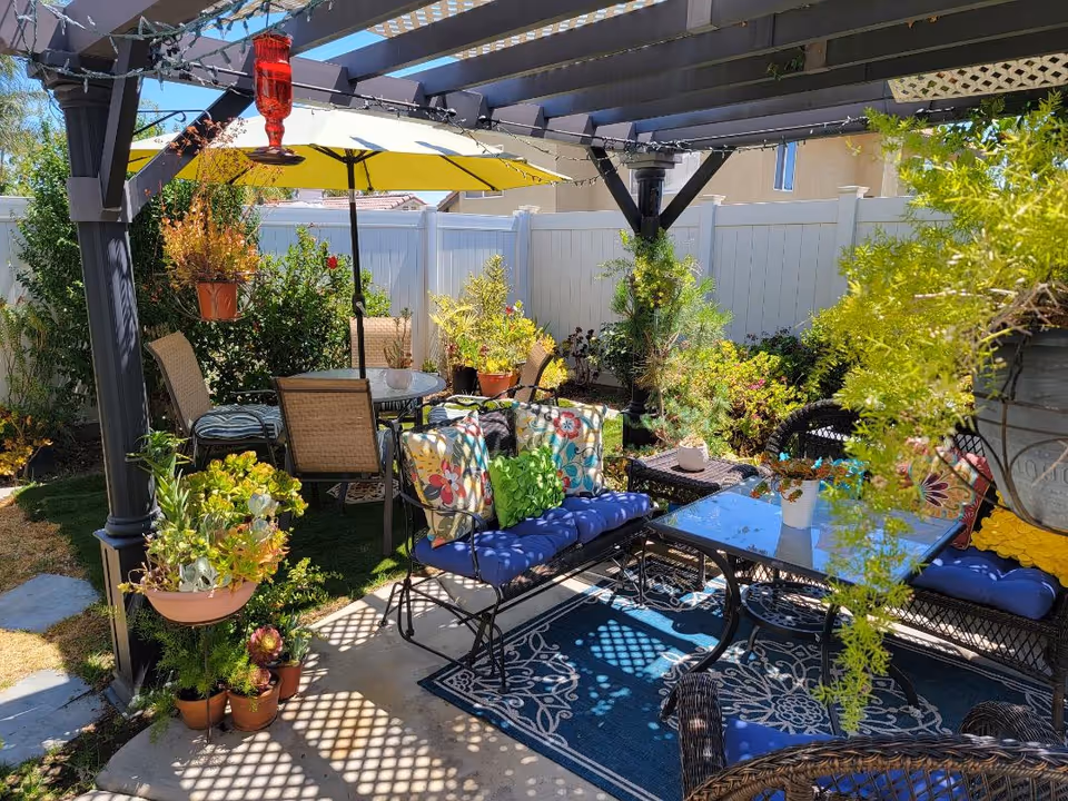 Outdoor patio area with a pergola overhead, decorated with string lights and hanging plants. There is a seating arrangement with cushioned chairs and a glass-top table on a patterned outdoor rug. A yellow umbrella shades a round table with chairs in the background, surrounded by various potted plants and greenery, enclosed by a white fence.
