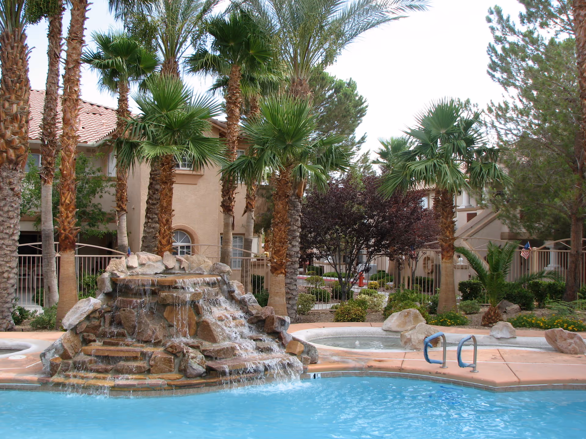 Outdoor swimming pool with a rock waterfall feature surrounded by palm trees and other greenery, with beige residential buildings in the background.