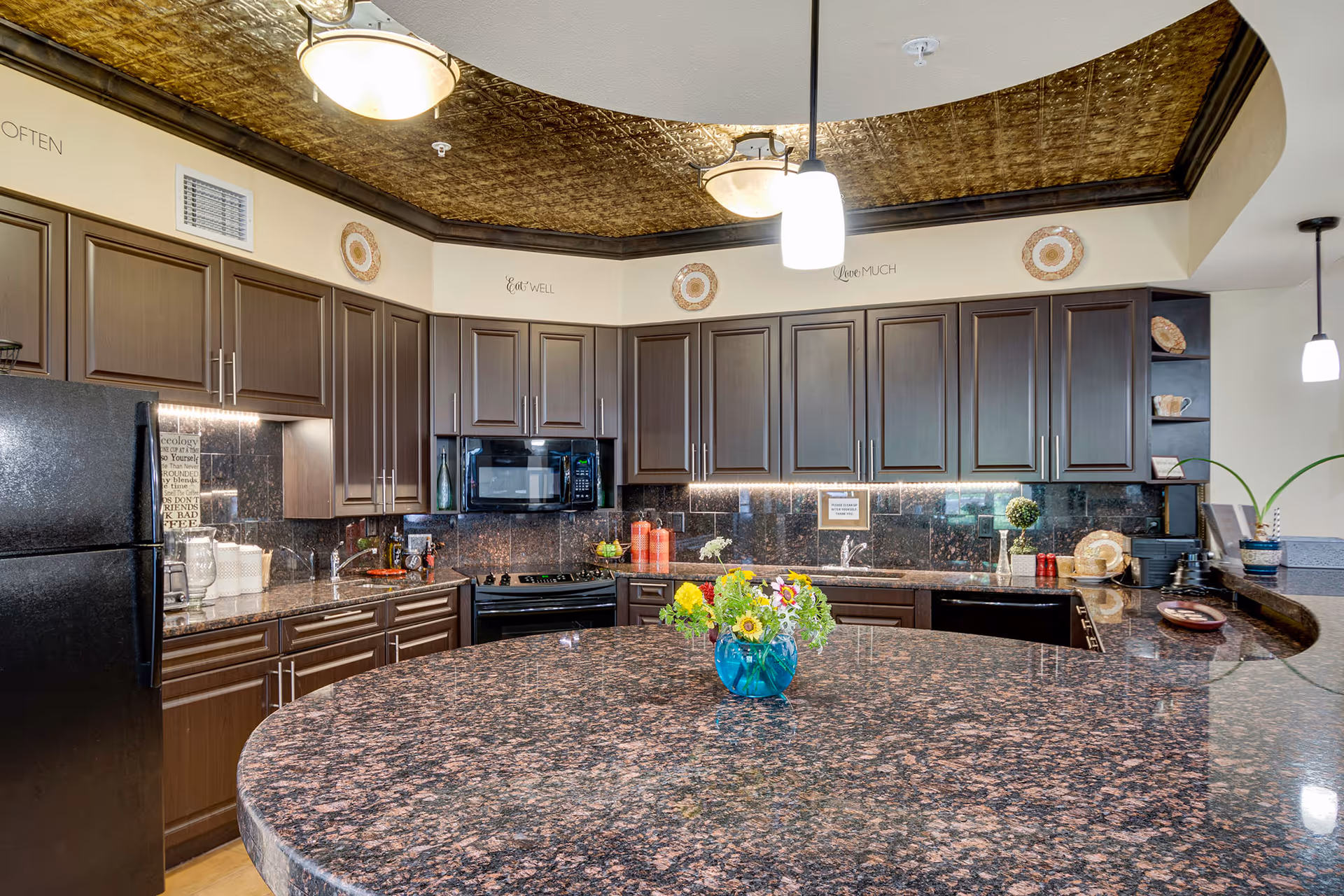 A modern kitchen with dark wood cabinets, black appliances including a refrigerator, microwave, and stove, and a large granite countertop island with a small blue vase holding colorful flowers. The ceiling has a decorative gold pattern with hanging light fixtures, and the walls have inspirational words like 'Eat Well' and 'Love Much'.