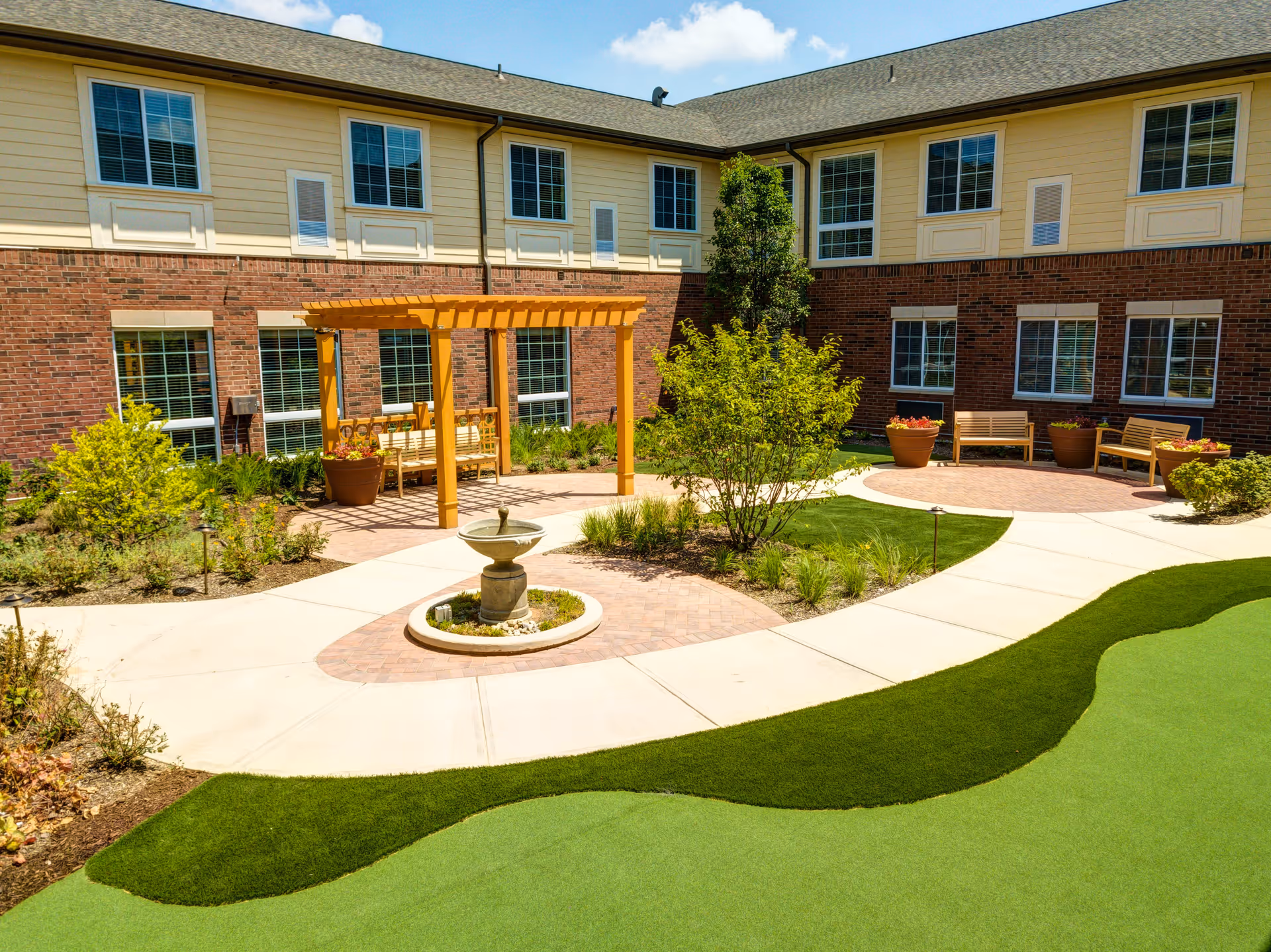 Outdoor courtyard area of a senior living facility with a small fountain in the center, surrounded by a paved walkway, green grass, shrubs, and a wooden pergola with benches underneath. The building has two stories with large windows and a combination of brick and beige siding.