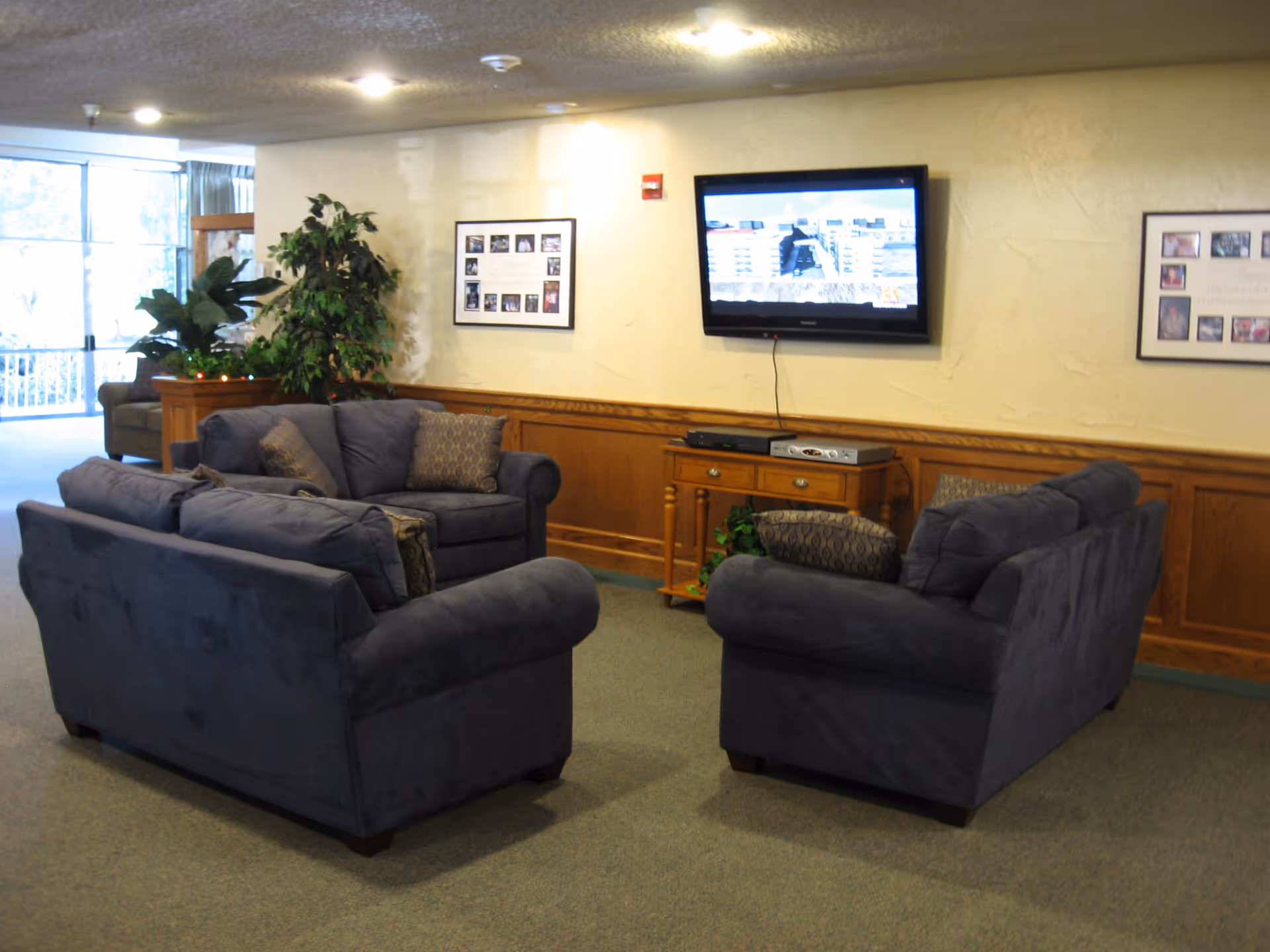 A lounge area with three blue sofas arranged facing a wall-mounted TV and a wooden console, with plants and framed photos on the walls.