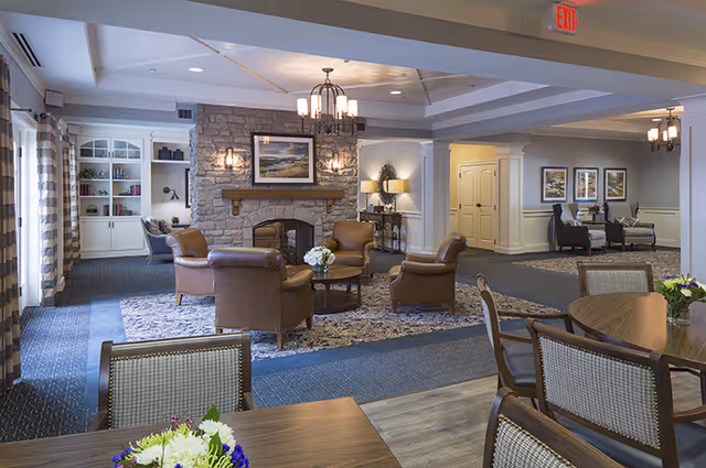 A spacious senior living common area with a stone fireplace, four brown leather armchairs arranged around a round coffee table with a flower centerpiece, and a chandelier overhead. The room has a mix of carpet and wood flooring, with additional seating areas including tables and chairs, built-in shelves with books, framed artwork on the walls, and soft lighting from lamps and wall sconces.
