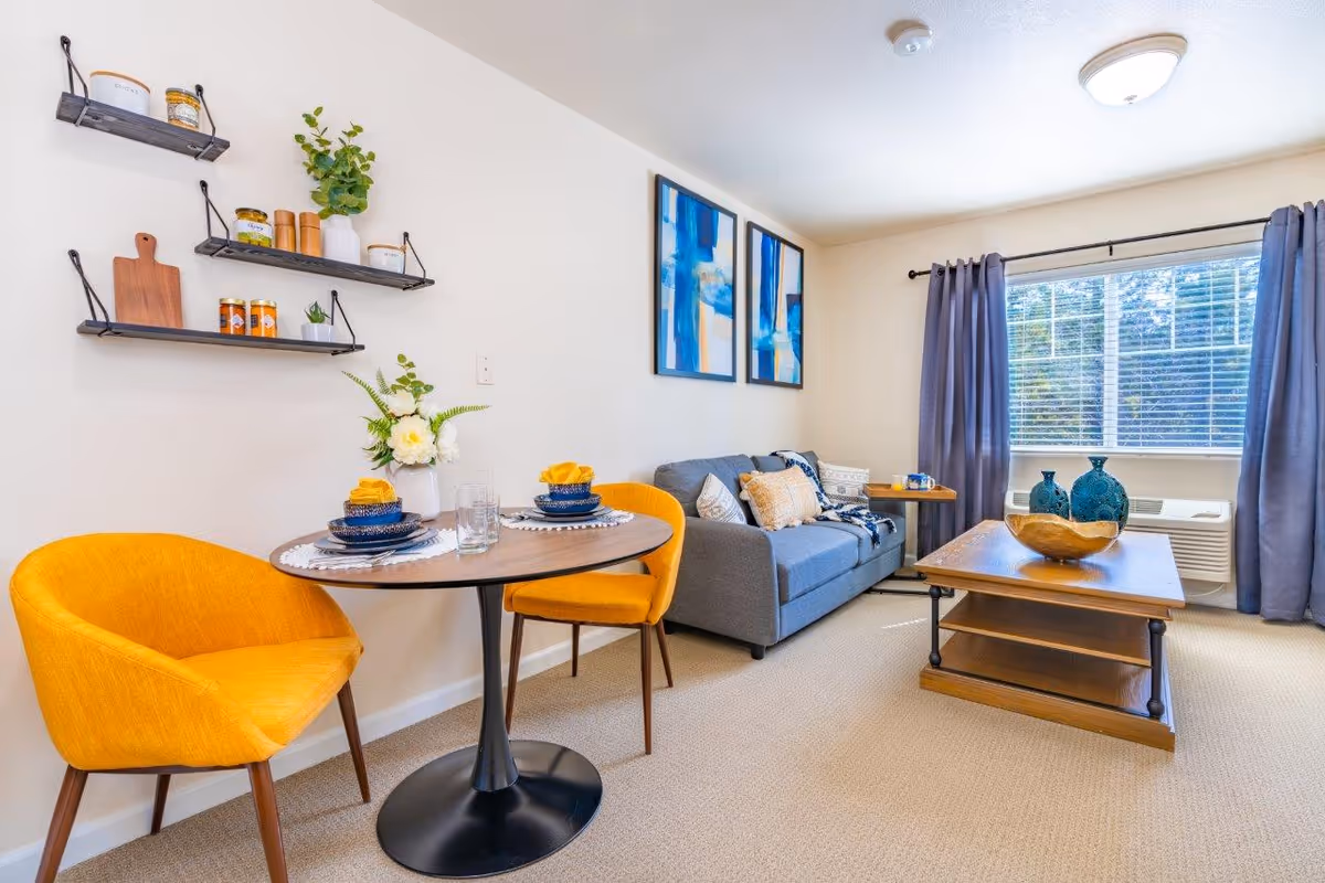 A cozy living room and dining area with a small round table set for two with yellow chairs. The room features a gray sofa with decorative pillows, a wooden coffee table with blue vases and a wooden bowl, two abstract blue paintings on the wall, and a large window with blue curtains letting in natural light.