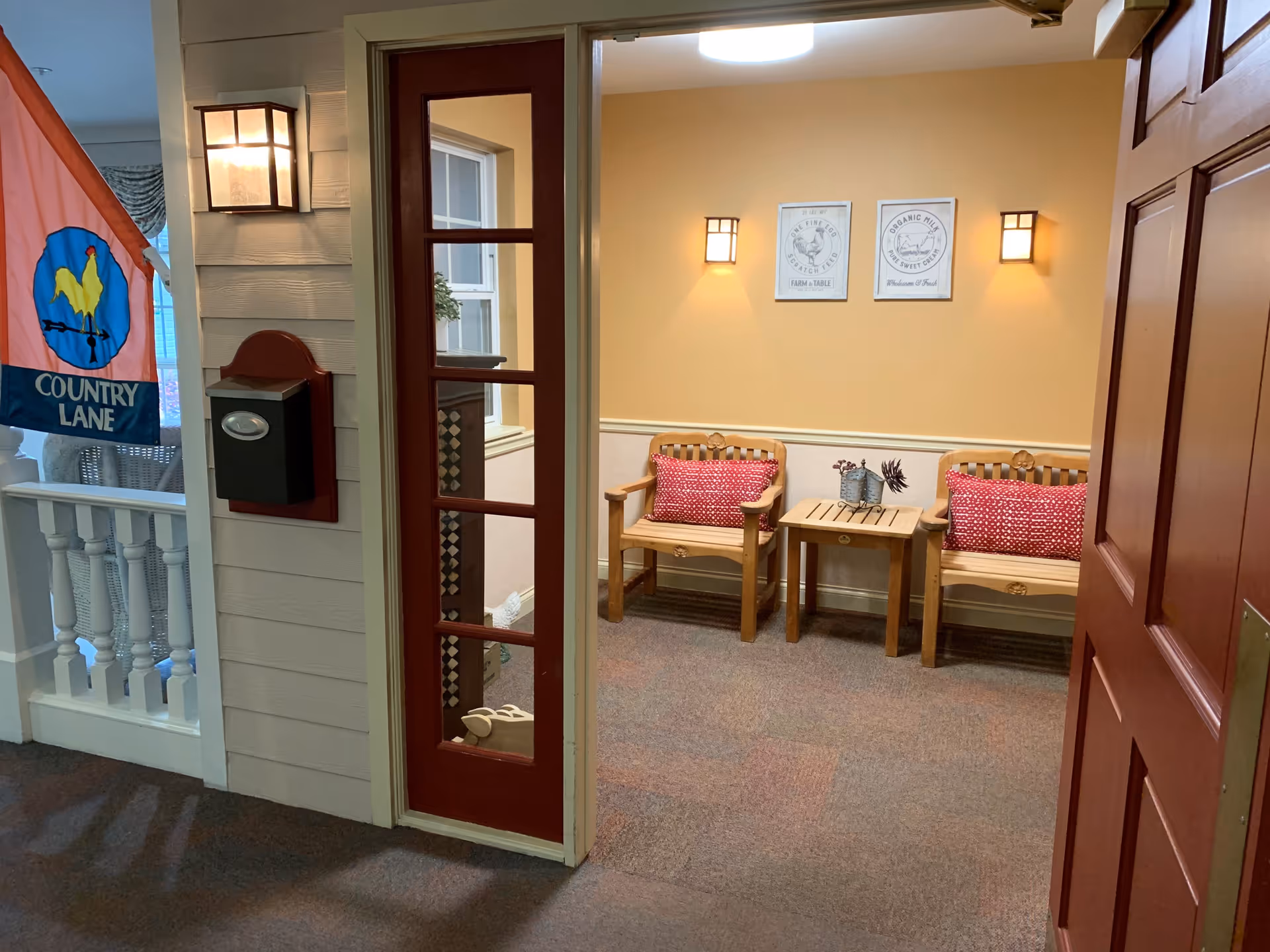 View through an open red door into a cozy seating area with two wooden chairs featuring red patterned cushions and a small wooden table between them. The walls are painted a warm beige color with two framed prints and two wall sconces providing soft lighting. To the left, there is a porch area with a white railing, a black mailbox, a wall-mounted light fixture, and a colorful flag with a rooster and the words 'Country Lane'.