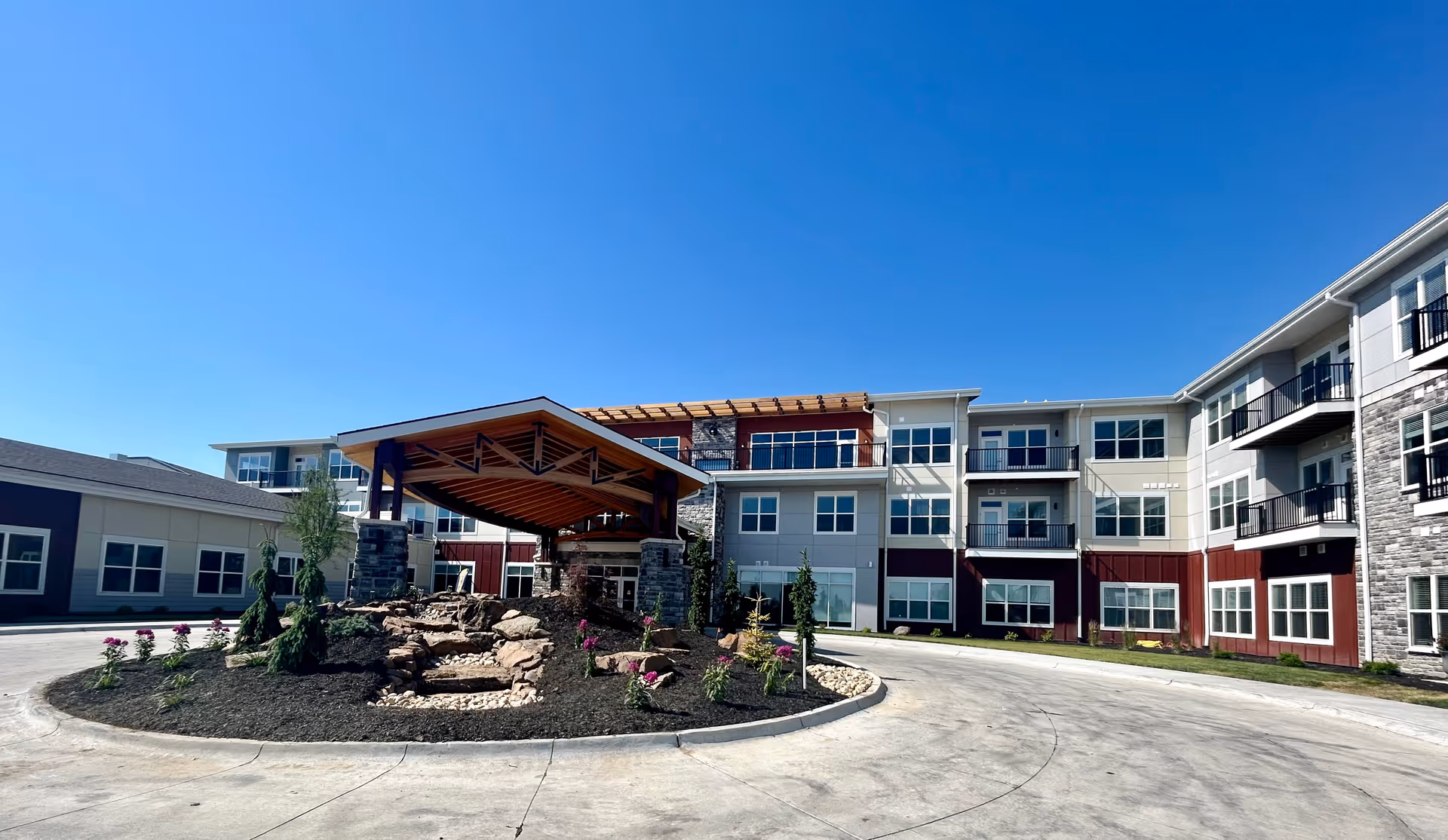 Exterior view of Hillcrest Highlands of Gretna, a multi-story senior living facility with a covered entrance, landscaped roundabout with rocks and flowers, and clear blue sky.