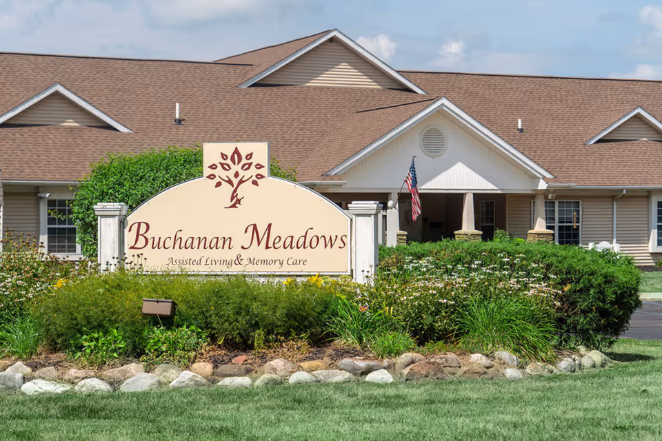 Exterior view of Buchanan Meadows assisted living and memory care facility with a large sign in front surrounded by greenery and flowers, and a building with a brown roof and beige siding in the background.