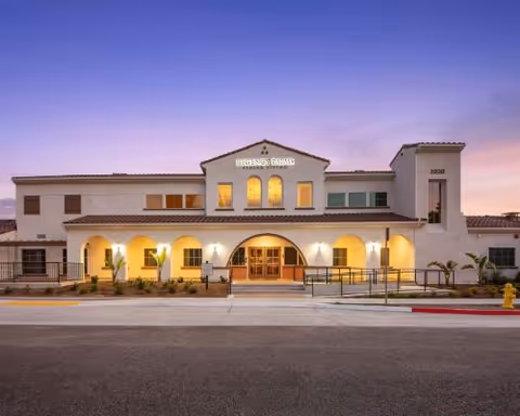 Front exterior view of Regency Palms Oxnard building at dusk with warm lights illuminating the entrance and arched windows, featuring a clear sky with a gradient from purple to light blue.