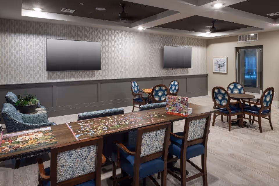 Community activity room with a long table holding a partially completed jigsaw puzzle, round tables and chairs, sofas, and wall-mounted TVs.