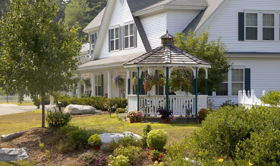 White multi-story building with a wooden gazebo and landscaped front yard with flowers and shrubs.