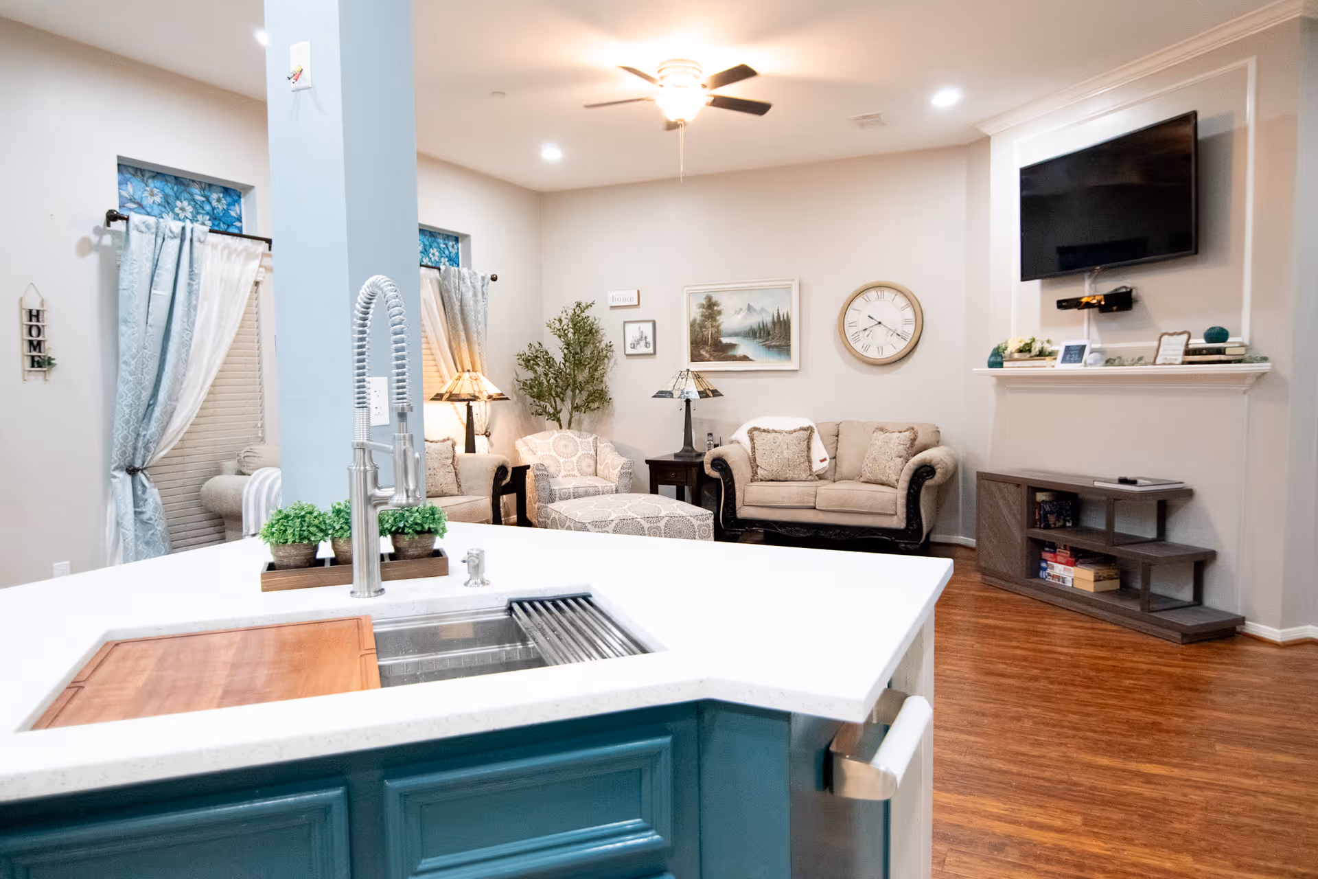 Bright and cozy living room area with beige sofas, a patterned armchair, a matching ottoman, and a wooden side table with a lamp. A large wall clock and a landscape painting hang on the wall. A flat-screen TV is mounted above a modern wooden entertainment unit with shelves holding books and decor. In the foreground, a kitchen island with a white countertop, a modern faucet, a sink, and small potted plants is visible. The room has wooden flooring and light-colored walls with windows dressed in light blue curtains.