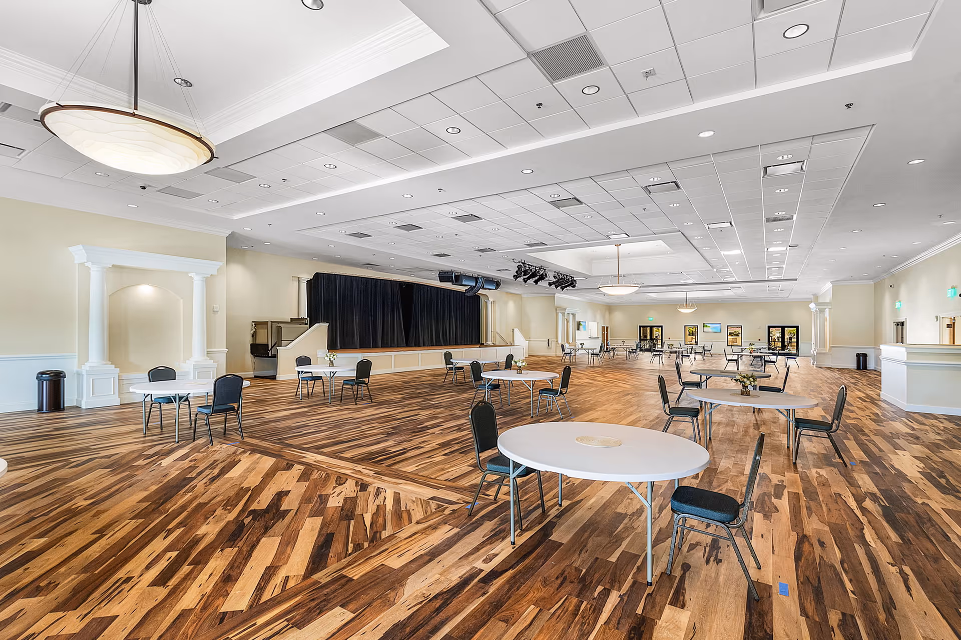 A large, spacious community room with wooden flooring and several round tables with chairs arranged around them. The room features a stage with black curtains at the far end, ceiling lights, and neutral-colored walls with white trim. The space appears well-lit and designed for gatherings or events.