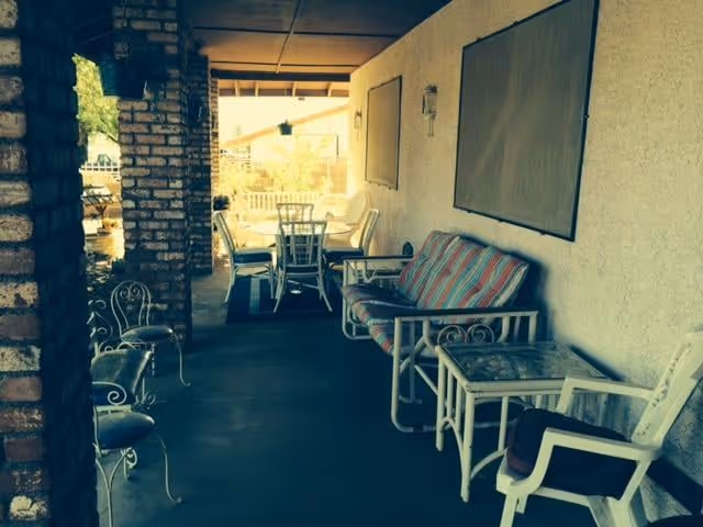 Covered outdoor patio area with a striped cushioned bench, white chairs, a glass-top side table, and a dining table with chairs in the background. The patio has brick columns and a concrete floor.