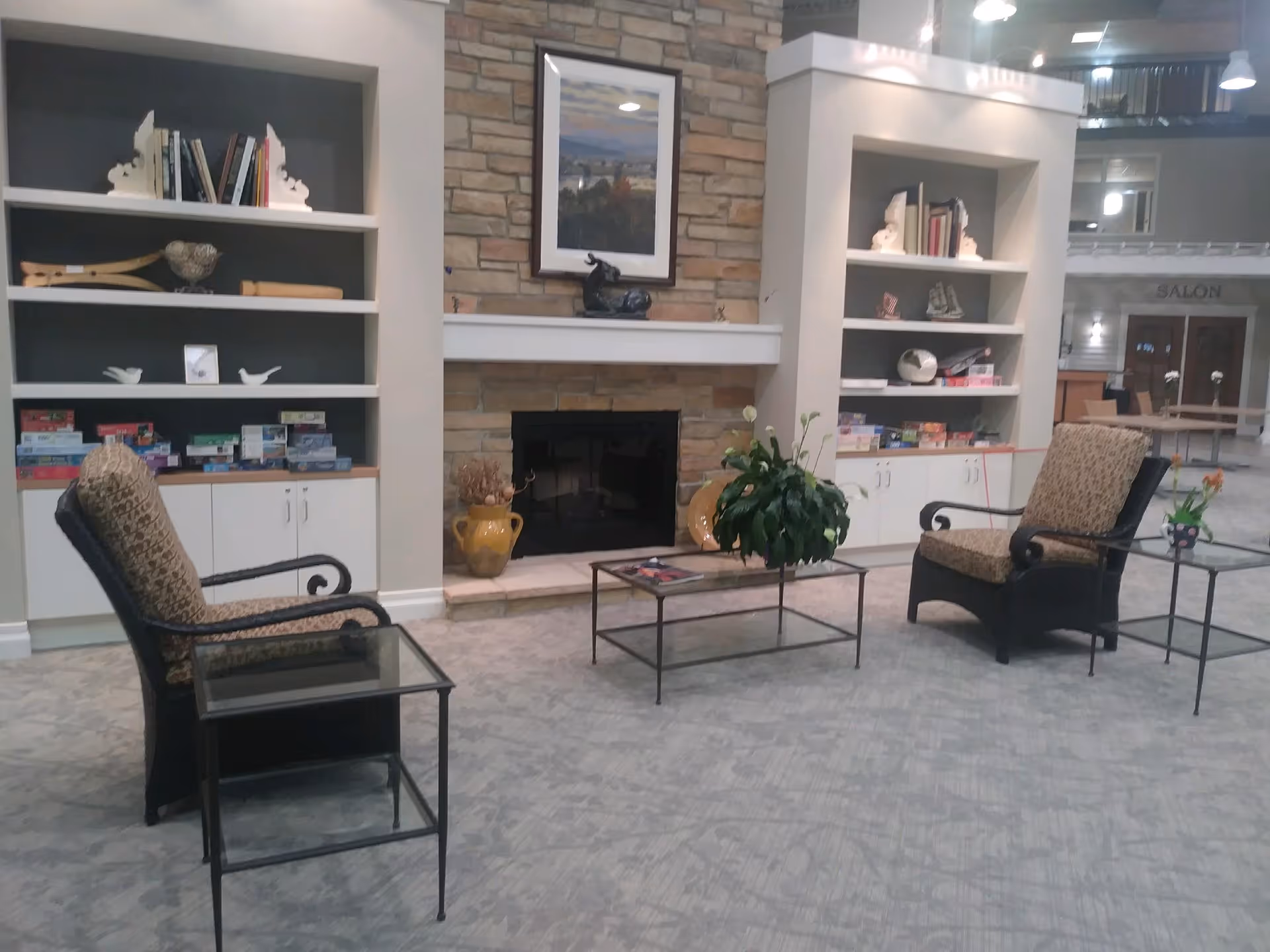 A cozy seating area in a senior living facility with two cushioned armchairs facing a stone fireplace. Built-in white shelves on either side of the fireplace hold books, decorative items, and board games. A glass coffee table with a green potted plant sits in front of the fireplace, and small side tables are next to each chair. The background shows a hallway with a sign for a salon.