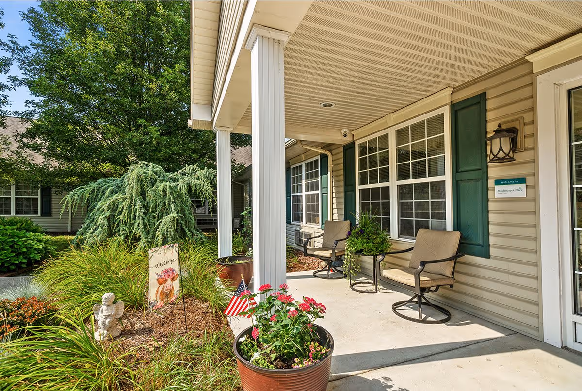 Covered porch area at Maidencreek Place with two cushioned chairs, a small table with a plant, and large windows with green shutters. The porch overlooks a garden with various plants, a small American flag, and a decorative welcome sign featuring a floral design. The building exterior is beige with white trim and a wall-mounted lantern light.