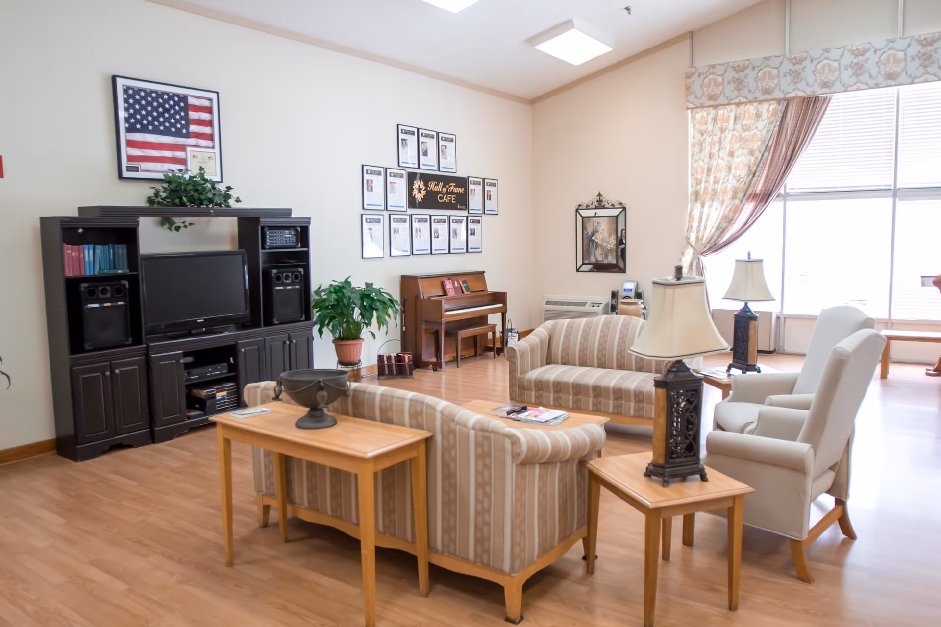 Communal living room with striped sofas and armchairs arranged around tables, a TV entertainment center, a piano, and large sunlit windows.