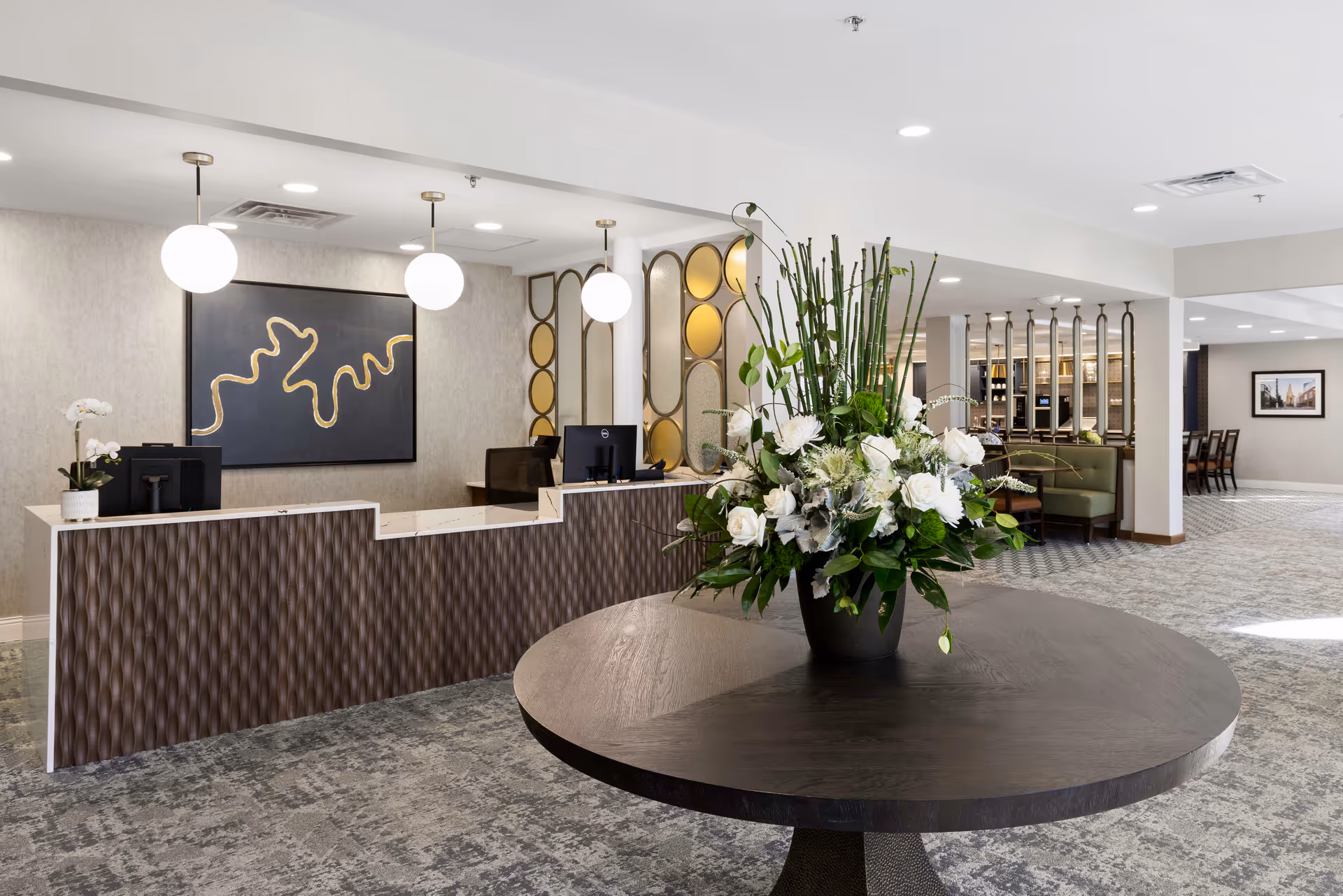 A modern senior living facility reception area with a curved front desk featuring two computer monitors. Behind the desk is a large abstract artwork with gold lines on a dark background. Three round pendant lights hang from the ceiling above the desk. In the foreground, there is a round wooden table with a large floral arrangement of white flowers and green foliage. The area has patterned carpet flooring and light-colored walls. To the right, there is a seating area with green cushioned benches and a dining area with tables and chairs visible in the background.
