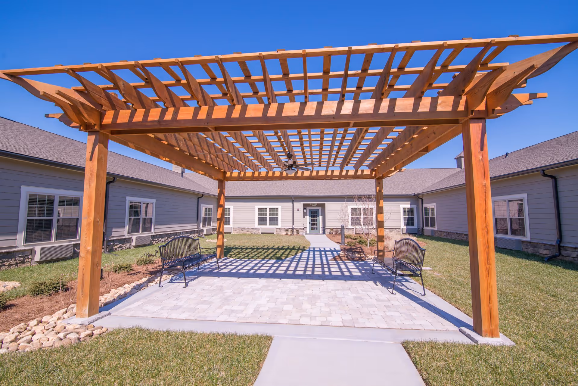 Outdoor courtyard area at Morning Pointe of Franklin featuring a wooden pergola with a ceiling fan, two black metal benches, paved stone flooring, and surrounding grass and landscaping. The courtyard is enclosed by a single-story building with multiple windows and a central door.