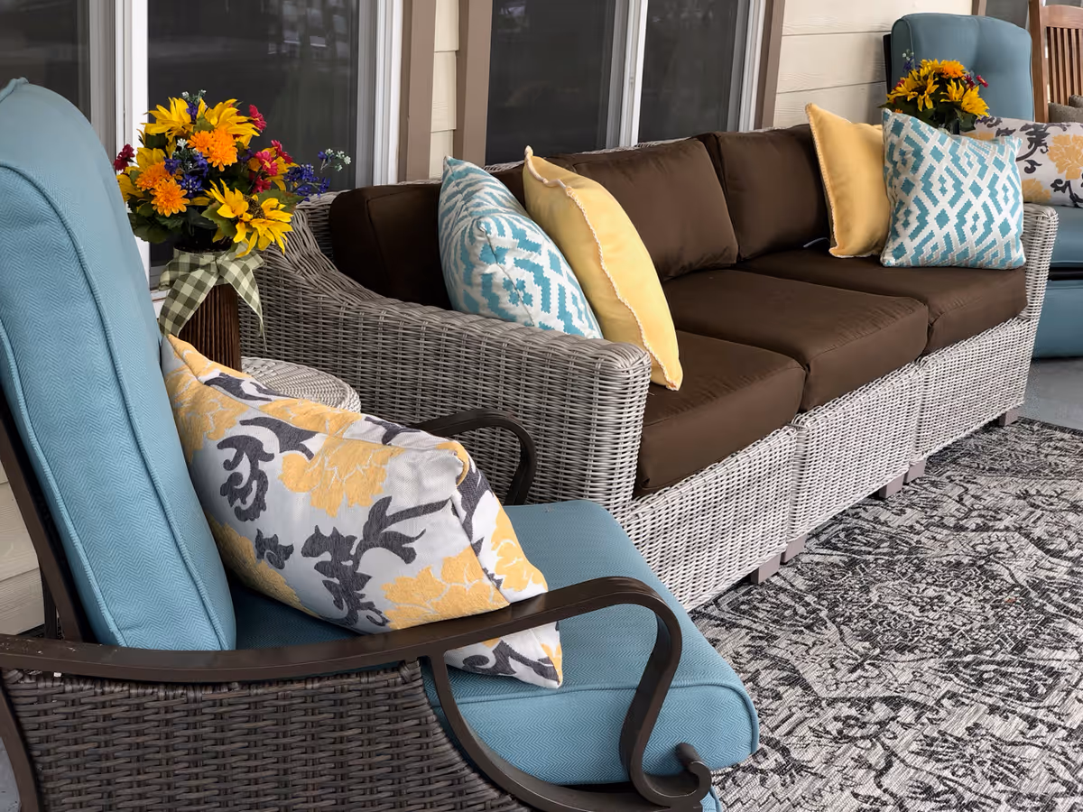 Outdoor patio seating area with wicker furniture including a brown cushioned sofa and blue cushioned chairs adorned with decorative pillows in yellow, blue, and floral patterns. There are two vases with colorful flowers placed behind the sofa, and a patterned rug on the floor.